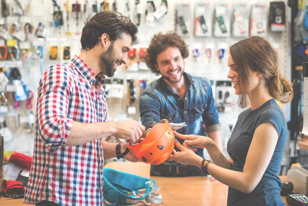 People engaging in a friendly retail interaction at an outdoor gear store, with a man handing an orange helmet to a woman, while another man smiles in the background, emphasizing high-touch customer service and relationship-building in luxury retail.